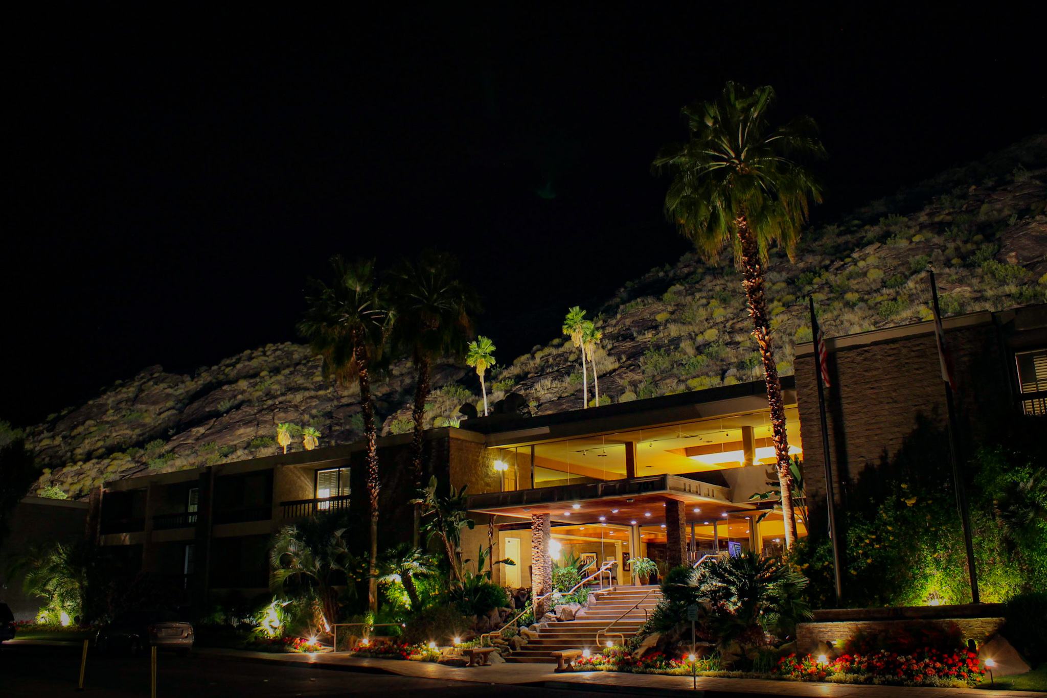 A well-lit hotel exterior at night with palm trees and a mountain in the background.