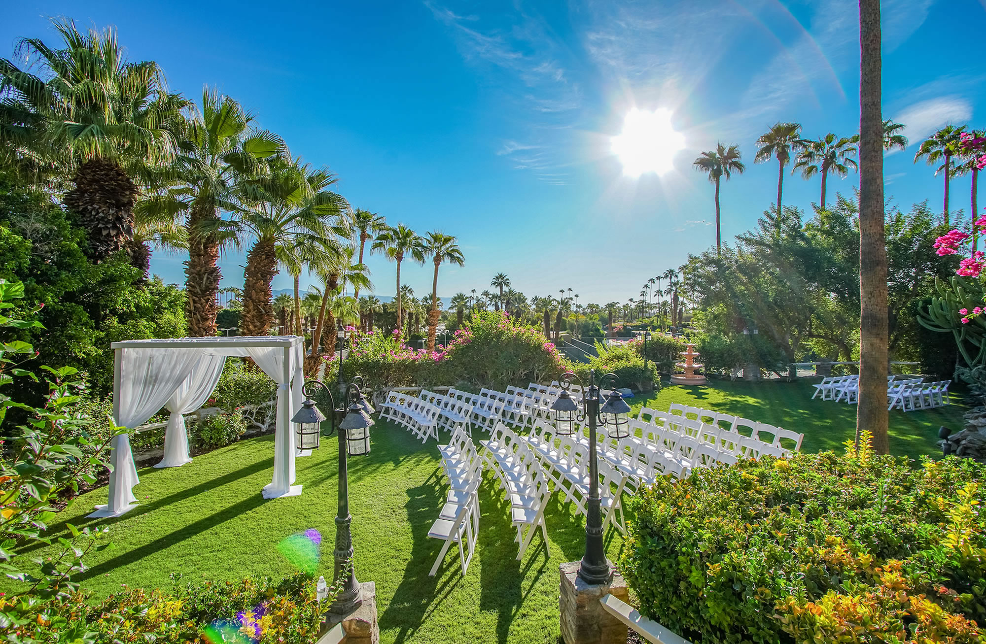 A lush outdoor event space set up for a ceremony with white chairs, an archway, and lanterns.
