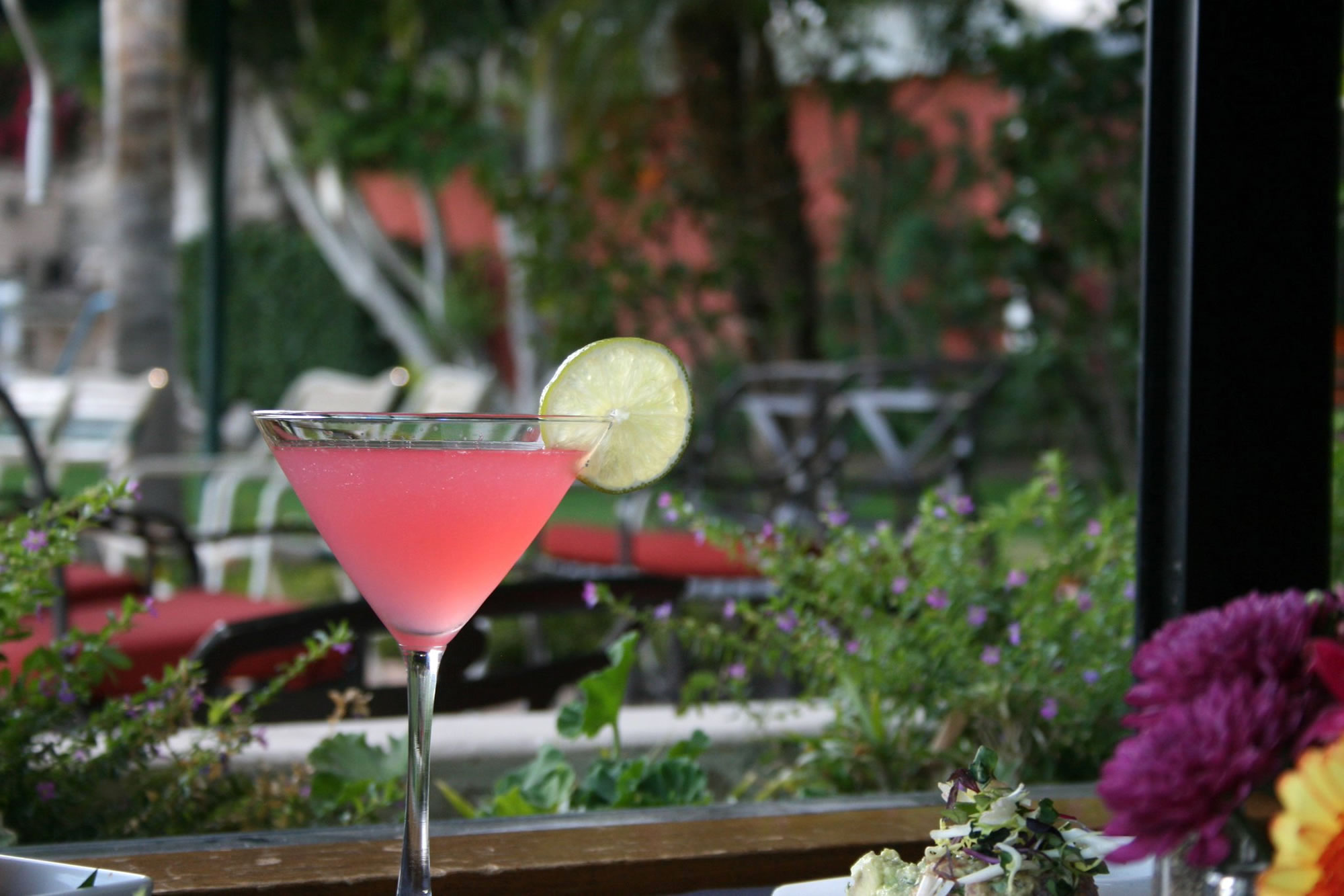 A pink cocktail in a martini glass with a lime wedge garnish, set against a blurred background of outdoor seating.